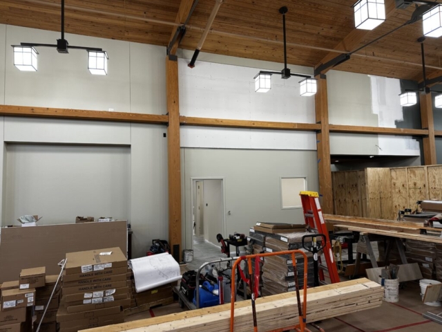 Interior of a commercial space under construction, showing exposed wooden beam ceiling with pendant lighting fixtures. Construction materials, cardboard boxes, a red ladder, and lumber are visible on the floor. The walls are painted light gray with wooden support beams.