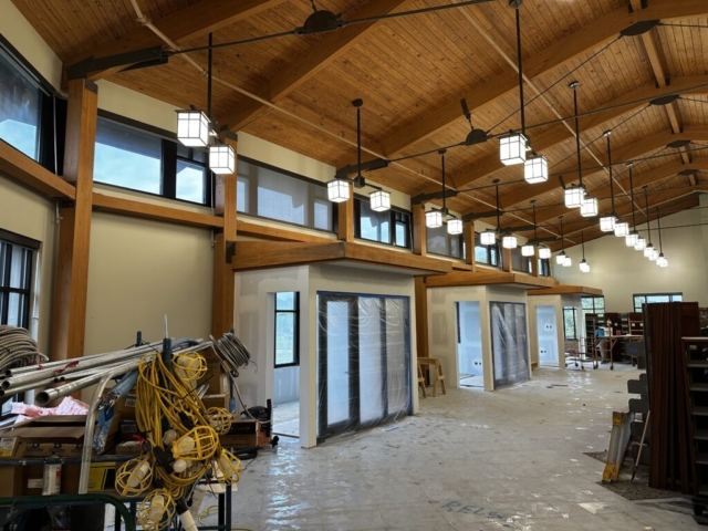 Large open commercial space with high vaulted wooden ceiling and clerestory windows. Multiple pendant lights hang in a row down the center. Several doorways or room divisions are covered in plastic sheeting. Construction equipment and materials are scattered on the concrete floor.