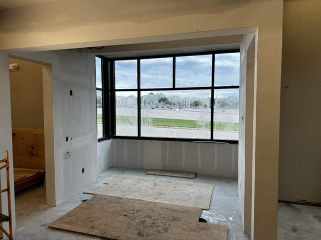 Unfinished window alcove or bay window area in a commercial building under construction. Large windows provide a view of a parking lot and trees outside. The floor is covered with plywood sheets and the walls are freshly drywalled but not yet finished. The space appears to be a future seating or display area.